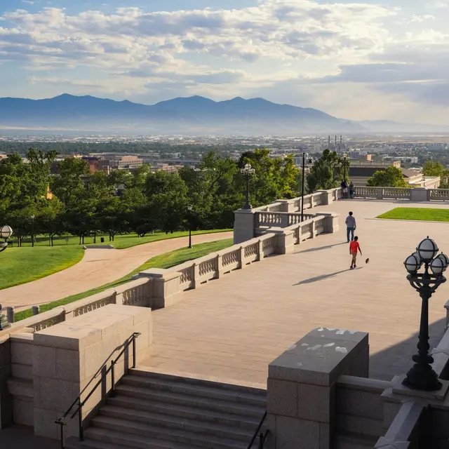 A scenic view from a park overlooking a city with mountains in the background. The sky is partly cloudy and the sun is shining, casting a warm light on the scene. A person is walking with a dog along a pathway, surrounded by lush green trees and well-maintained lawns.