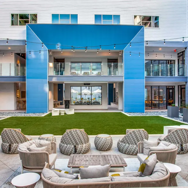 A modern apartment complex courtyard featuring a green lawn, comfortable seating arrangements, and blue accents on the building facade.