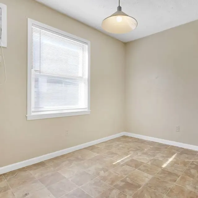 A spacious empty room with beige walls and tile flooring. There is a window with blinds letting in natural light, and a ceiling light fixture provides additional illumination. An air conditioning unit is mounted on the wall.