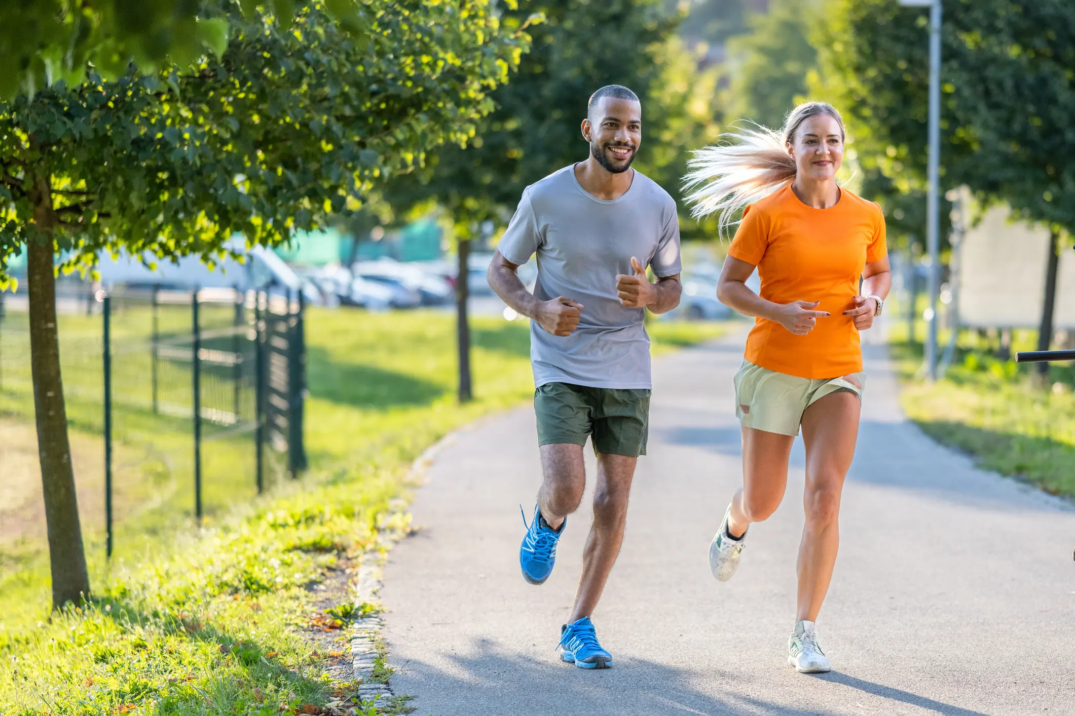 Two people jogging together in a park. One man is wearing a light grey shirt and shorts, while a woman is dressed in an orange shirt and light shorts. They are smiling and appear to be enjoying their run amidst green trees.