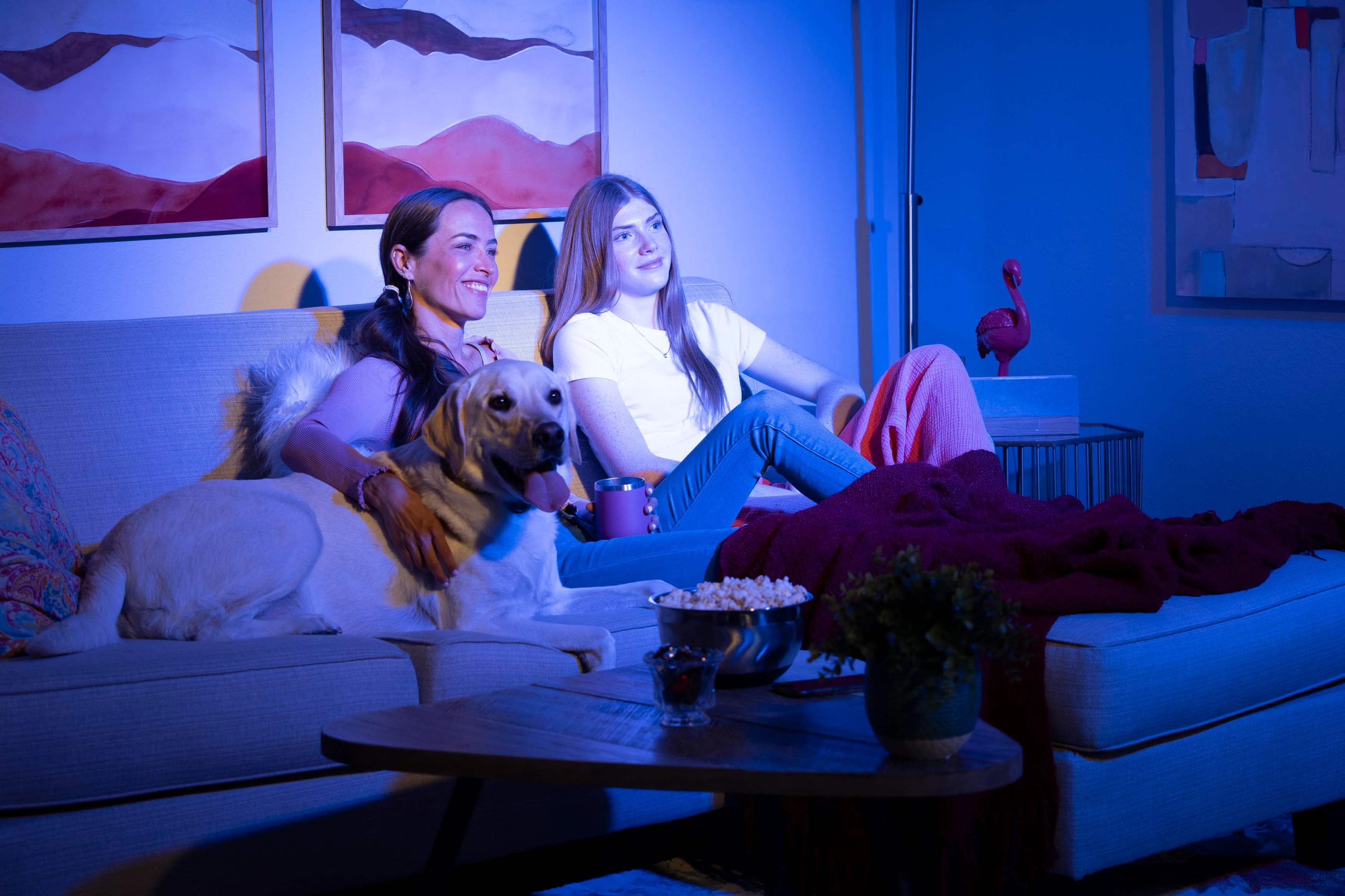 Two women sitting on a sofa with a dog, enjoying a cozy evening together. The room is illuminated with soft blue light.