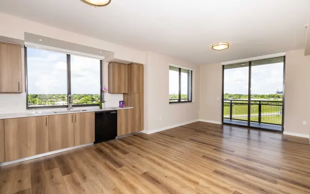 A spacious modern apartment interior featuring a kitchen area with wooden cabinets and a black dishwasher, an open living area with wooden flooring, large windows allowing plenty of natural light, and a view of greenery outside.