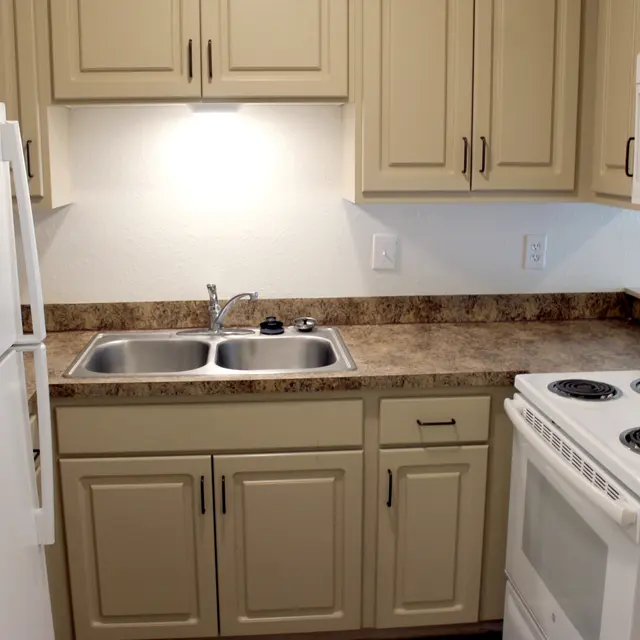 Compact Modern Kitchen A small kitchen featuring beige cabinets, a double basin stainless steel sink, a white refrigerator, and a white stove with an oven. The countertop is brown with a subtle pattern, and there is a light above the sink illuminating the area.