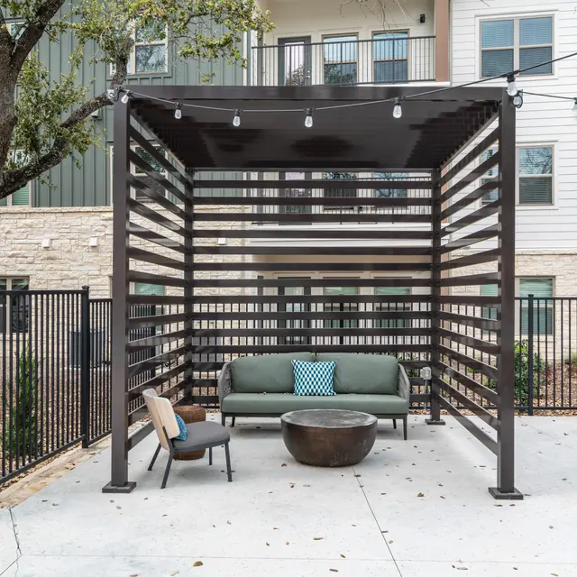 An outdoor lounge area with a pergola, containing a green sofa with a blue cushion, a round coffee table, and a single chair. Surrounding elements include trees and an apartment building in the background.