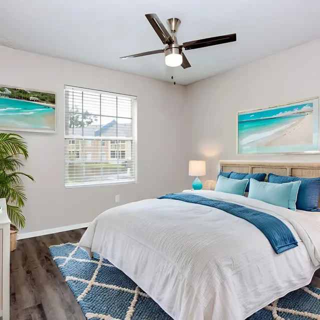 A modern bedroom featuring a large bed with white bedding and teal accents, two bedside lamps, and beach-themed artwork on the walls. There's a television on a white stand and a green plant in the corner, with a geometric area rug on the floor.