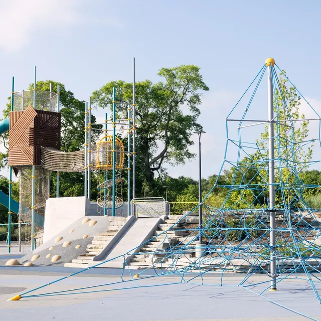 An outdoor playground featuring various play structures. On the left, a large slide and climbing area made of wooden materials, and on the right, a colorful rope climbing structure against a clear blue sky. There are trees in the background and children playing nearby.