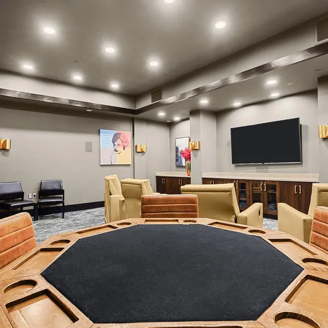 A modern game room with seating and a large poker table in the center. There are black chairs, a television on the wall, and a popcorn machine in the corner. Decorative pieces include artwork and flowers.