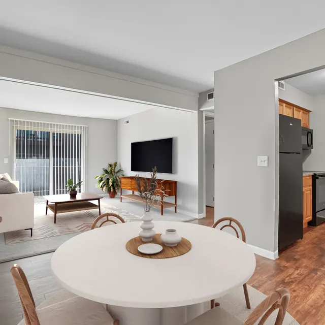A modern apartment interior featuring a dining table set in the foreground, a living area with a large TV and sofa in the background, and a kitchen visible to the right.