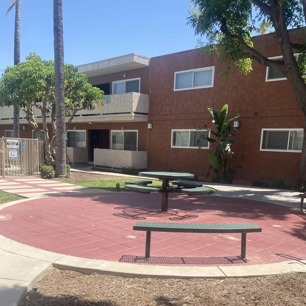 A small courtyard area with benches and a picnic table, surrounded by an apartment complex and palm trees under clear blue skies.