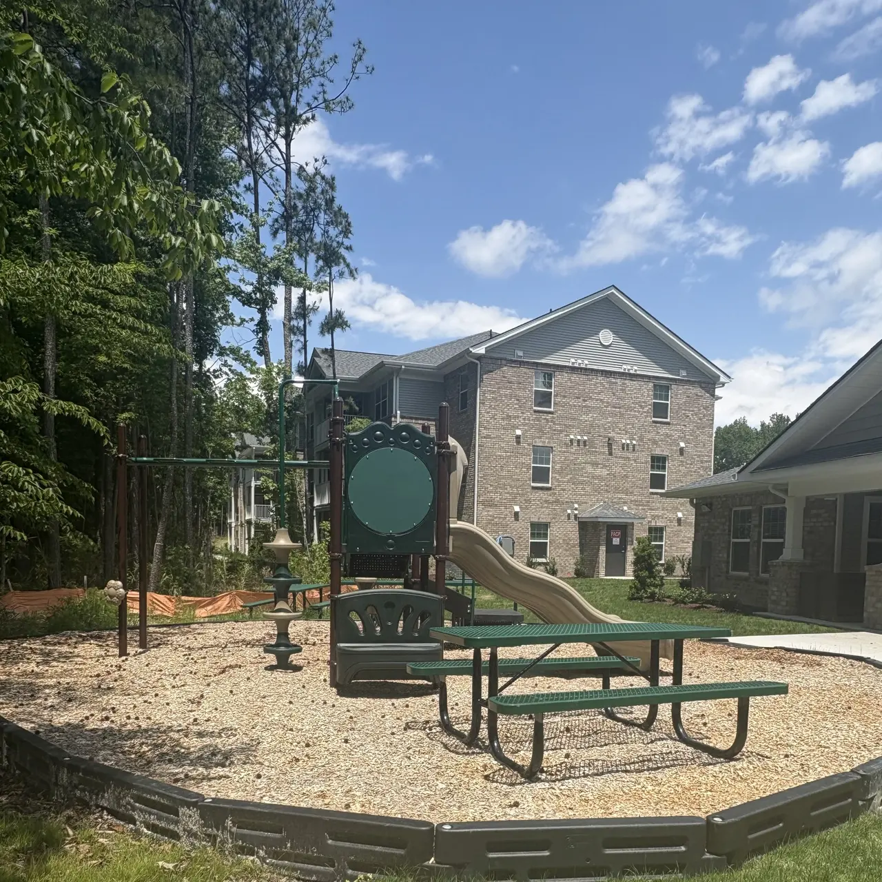 A small playground area with a swing set, slide, and benches surrounded by trees and a residential building in the background. Bright blue sky with some clouds is visible.