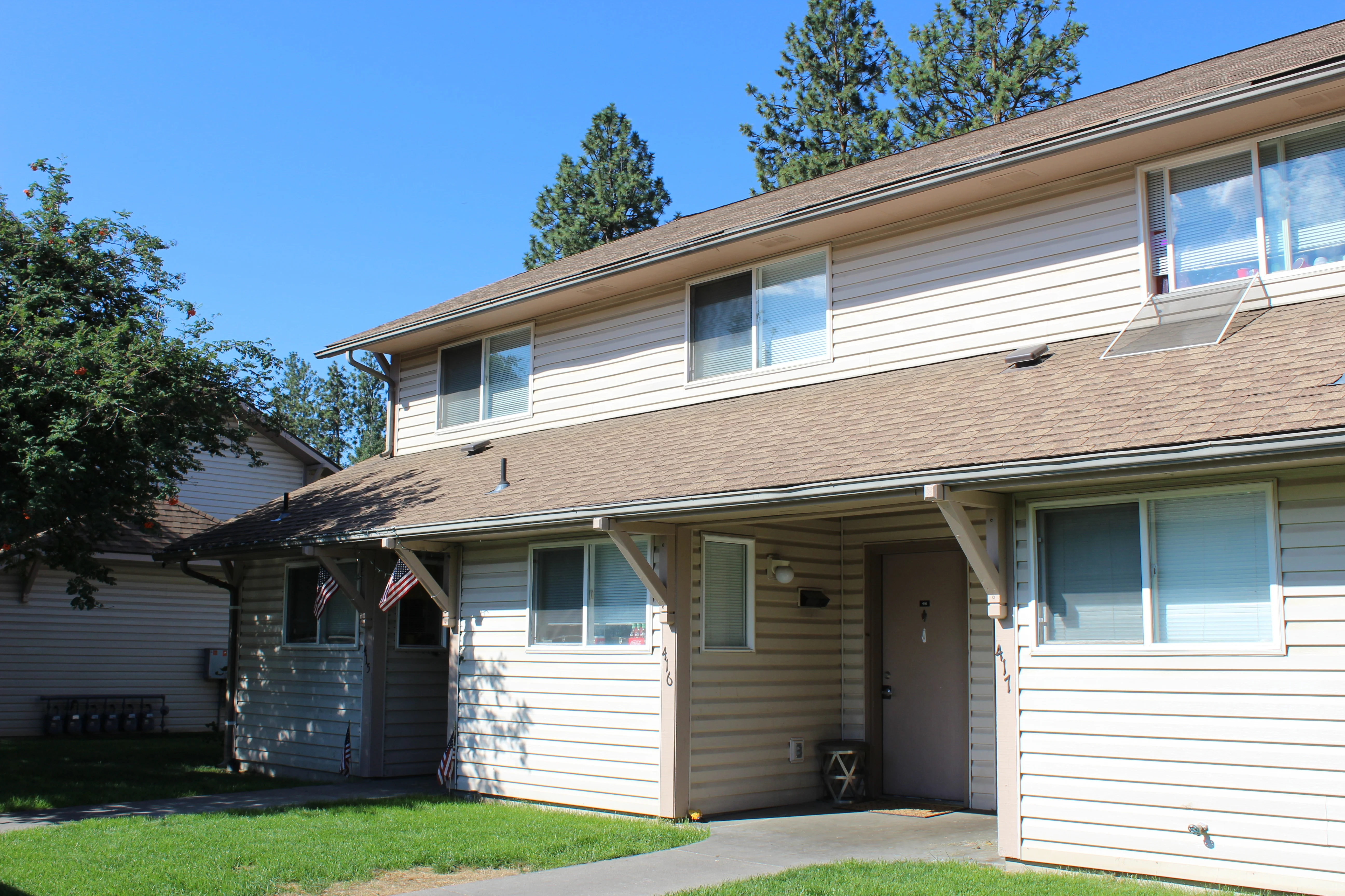 Exterior view of a two-story apartment building with a walkway, green lawn, and trees in the background.