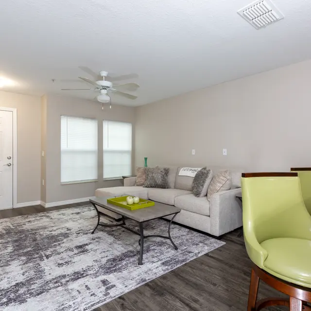A modern living room featuring a light-colored sofa, a coffee table with a bowl of green apples, and unique green chairs. The walls are painted light beige, and the flooring is dark wood.