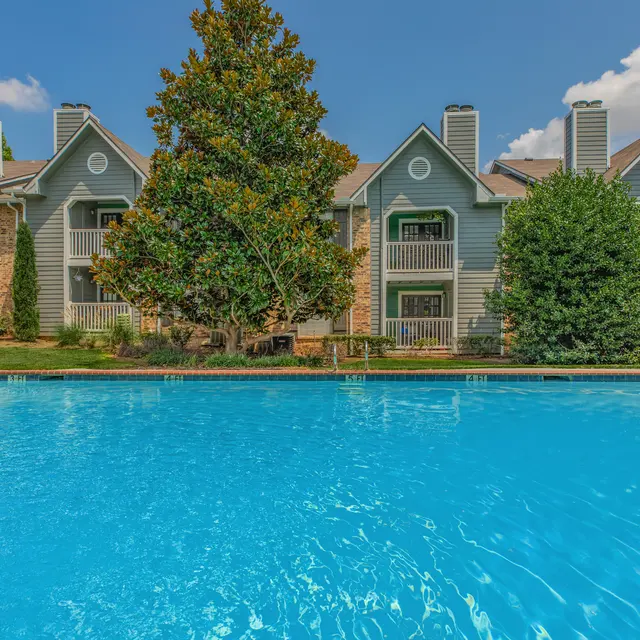 A scenic view of an apartment complex featuring a blue swimming pool in the foreground, with several trees and landscaped areas. The sky is clear with a few clouds, and the buildings have a light-colored exterior with balconies.