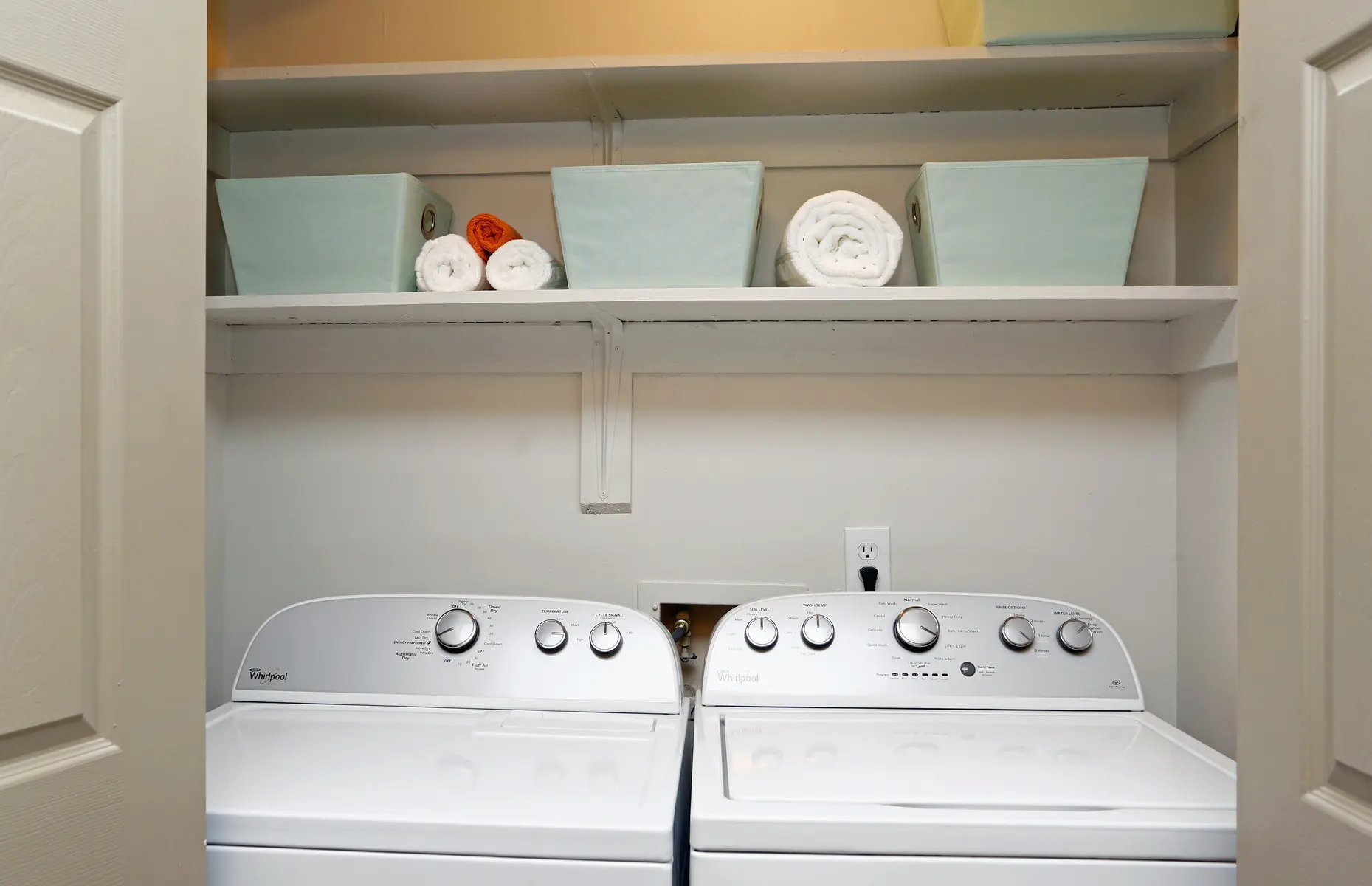 A compact laundry area featuring a washer and dryer side by side, with shelves above holding several colorful and white rolled towels, along with blue storage bins.