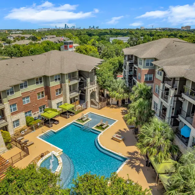 Aerial view of a luxury apartment complex featuring a swimming pool surrounded by lounge chairs and palm trees, with a city skyline in the background.