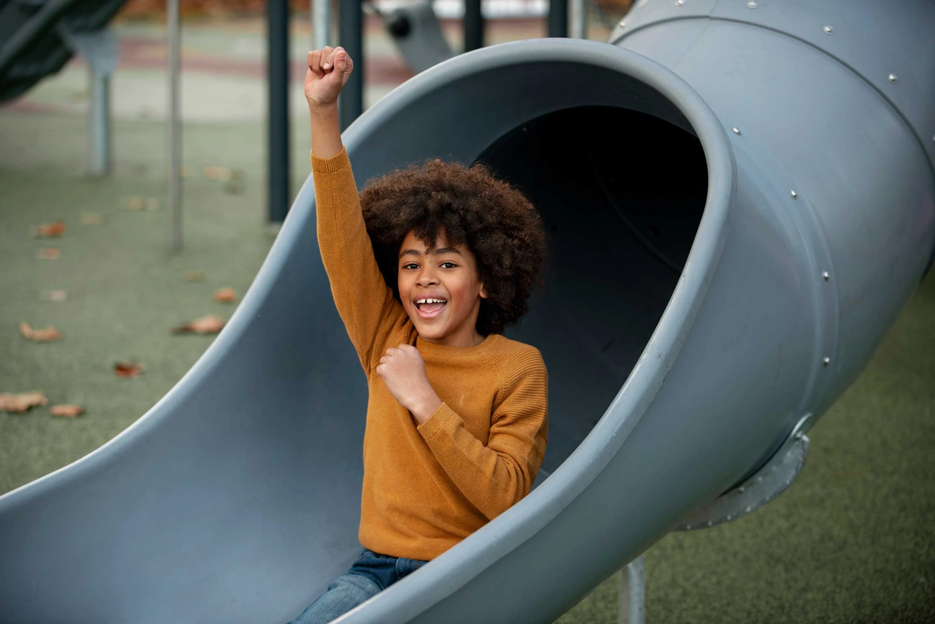 A joyful child with curly hair sits inside a gray slide, raising one fist in celebration. The playground setting has greenery and structures in the background.