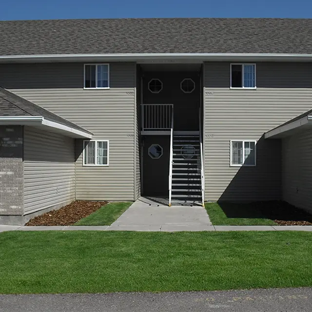 Two-Story Apartment Building Exterior Exterior view of a two-story apartment building with a staircase at the center, flanked by two garages on either side, surrounded by green grass and a clear blue sky.