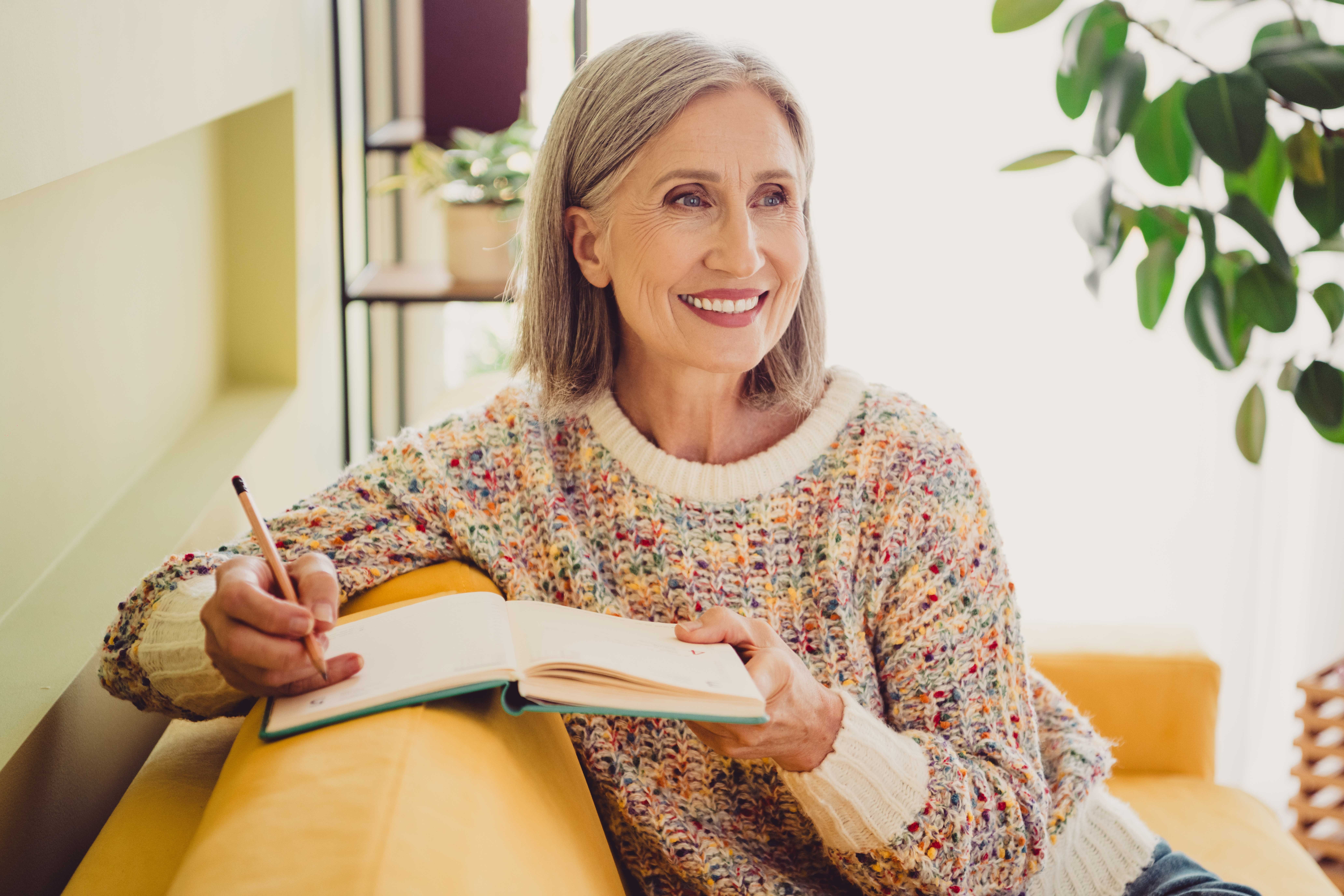 A smiling senior woman in a colorful sweater writing in a notebook while sitting on a yellow sofa.