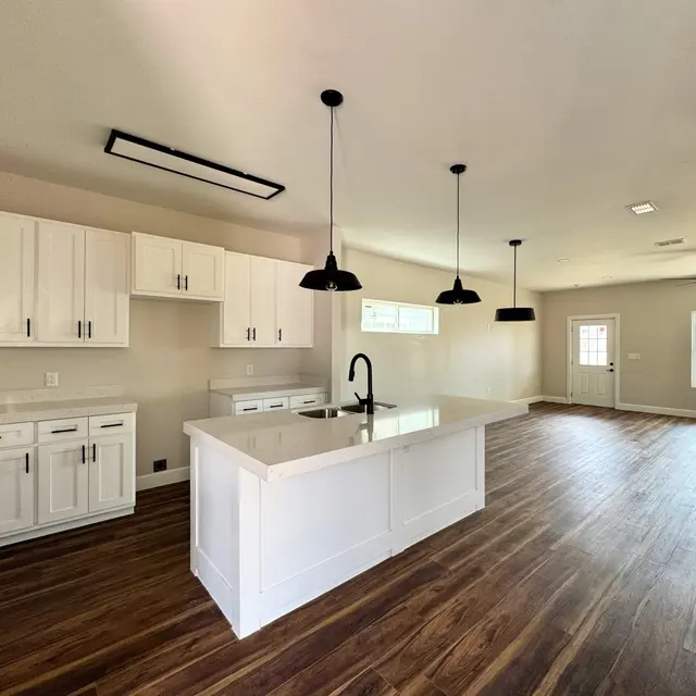 Spacious modern kitchen featuring white cabinetry, a large island with a sink, and dark pendant lighting. Hardwood flooring runs throughout the open space leading to a living area with natural light coming through large windows.