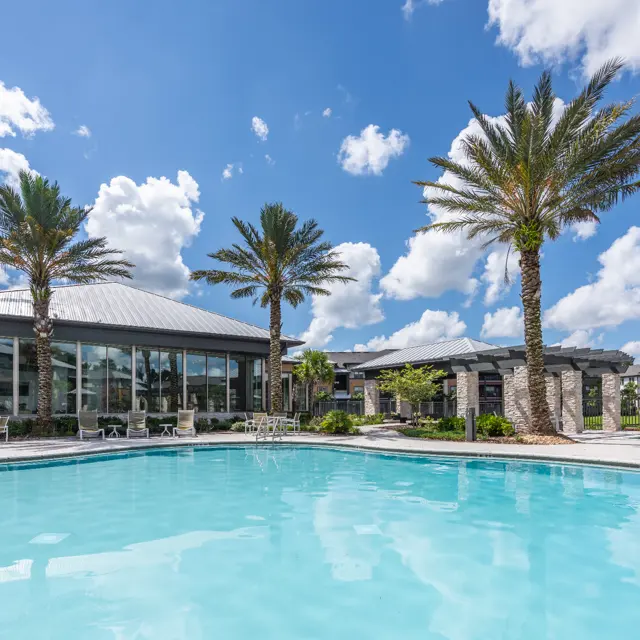 A swimming pool surrounded by palm trees and a modern building under a blue sky with fluffy clouds.