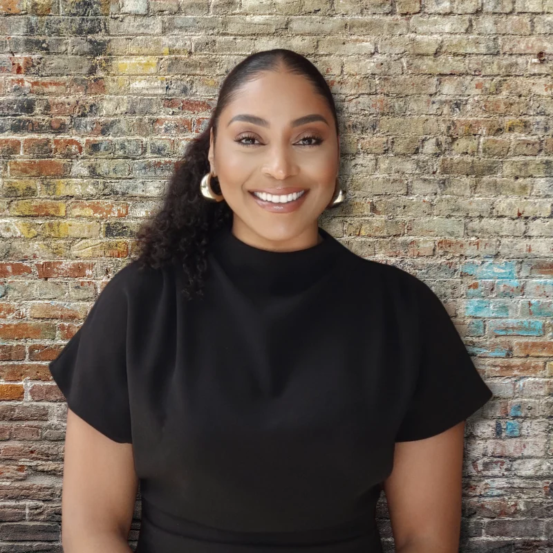 Portrait of a Smiling Woman in Black A woman smiling, wearing a black t-shirt, with her hair styled partially up and earrings, standing in front of a textured brick wall.