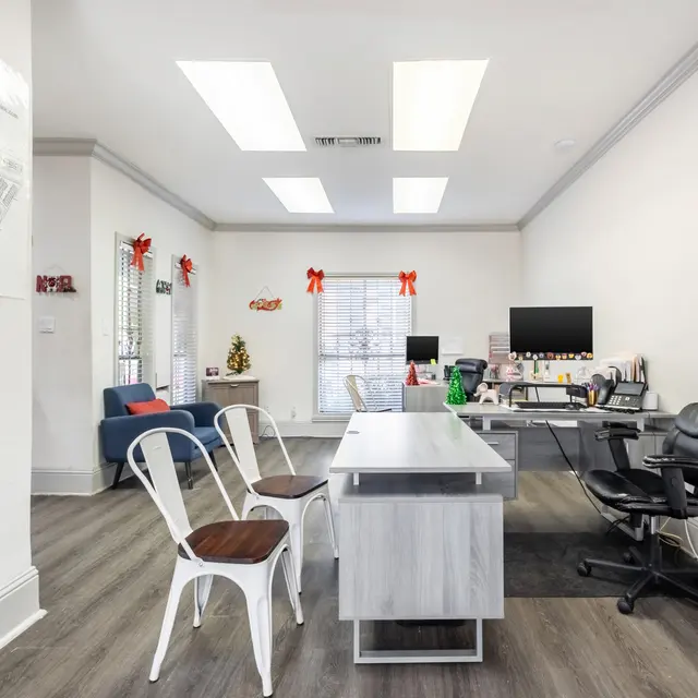 A modern office interior featuring a central desk, a black office chair, and white chairs. The walls are white with a map on one side and seasonal decorations. There's a window with natural light coming in.