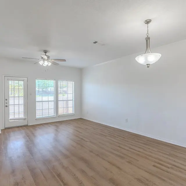 Spacious living room with wooden flooring, white walls, and a ceiling fan. The room includes a fireplace and large windows with sliding doors leading outside.