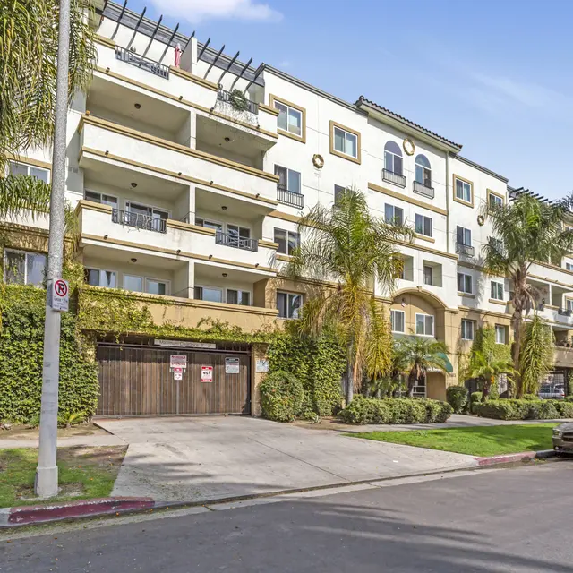 A modern apartment building with multiple floors, featuring balconies and palm trees in the foreground, located on a sunny street.