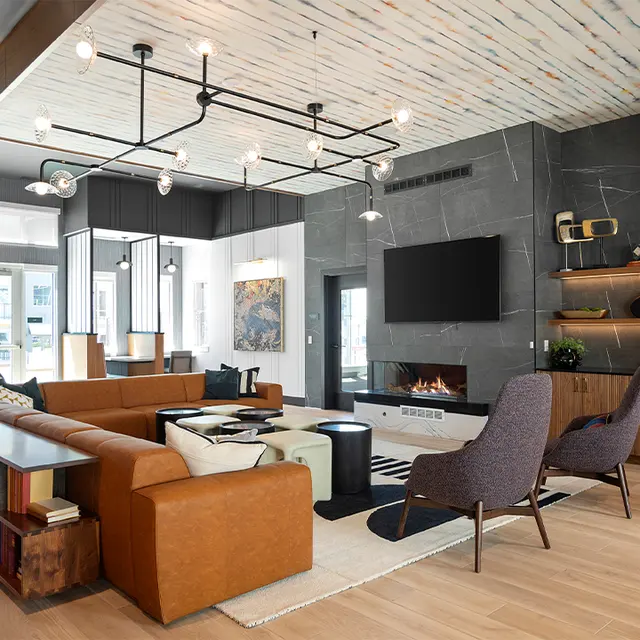 A modern living room with a sectional sofa, accent chairs, and a coffee table. The wall features a large TV and wooden shelving. Large windows provide natural light.