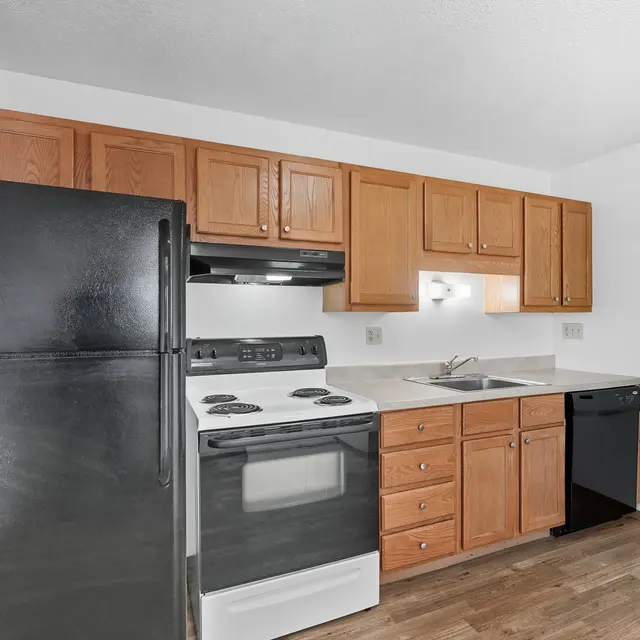 A modern kitchen featuring oak cabinets, a black refrigerator, a black dishwasher, an electric stove, and a stainless steel sink. The floor is wooden and the walls are white.