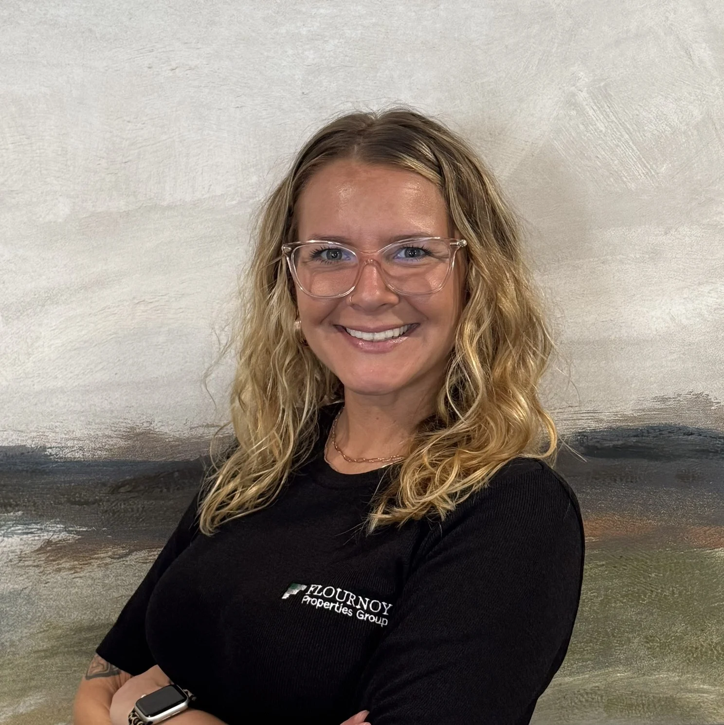 Professional Portrait A smiling young woman with curly blonde hair wearing glasses and a black shirt with a logo, standing with her arms crossed in front of an abstract background.