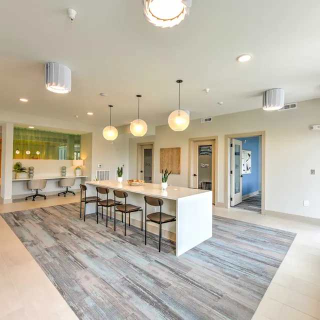 A spacious common area in a modern apartment building featuring a long table with chairs, pendant lights, and a decorative rug. The walls are painted in light colors with large windows providing natural light.