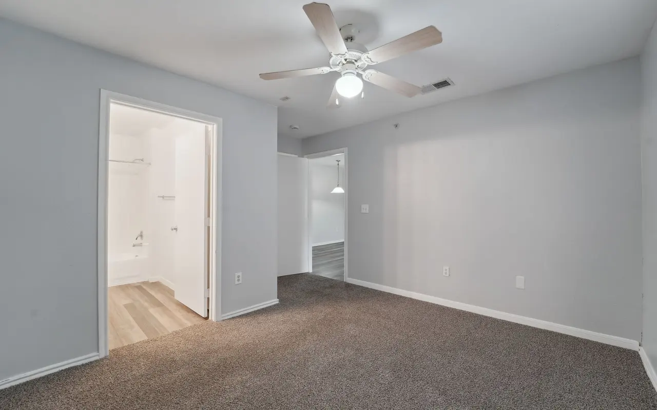 A spacious room with light gray walls and carpet flooring, featuring a ceiling fan and an open layout. Two doorways lead to adjoining spaces, one of which leads to a bathroom area.