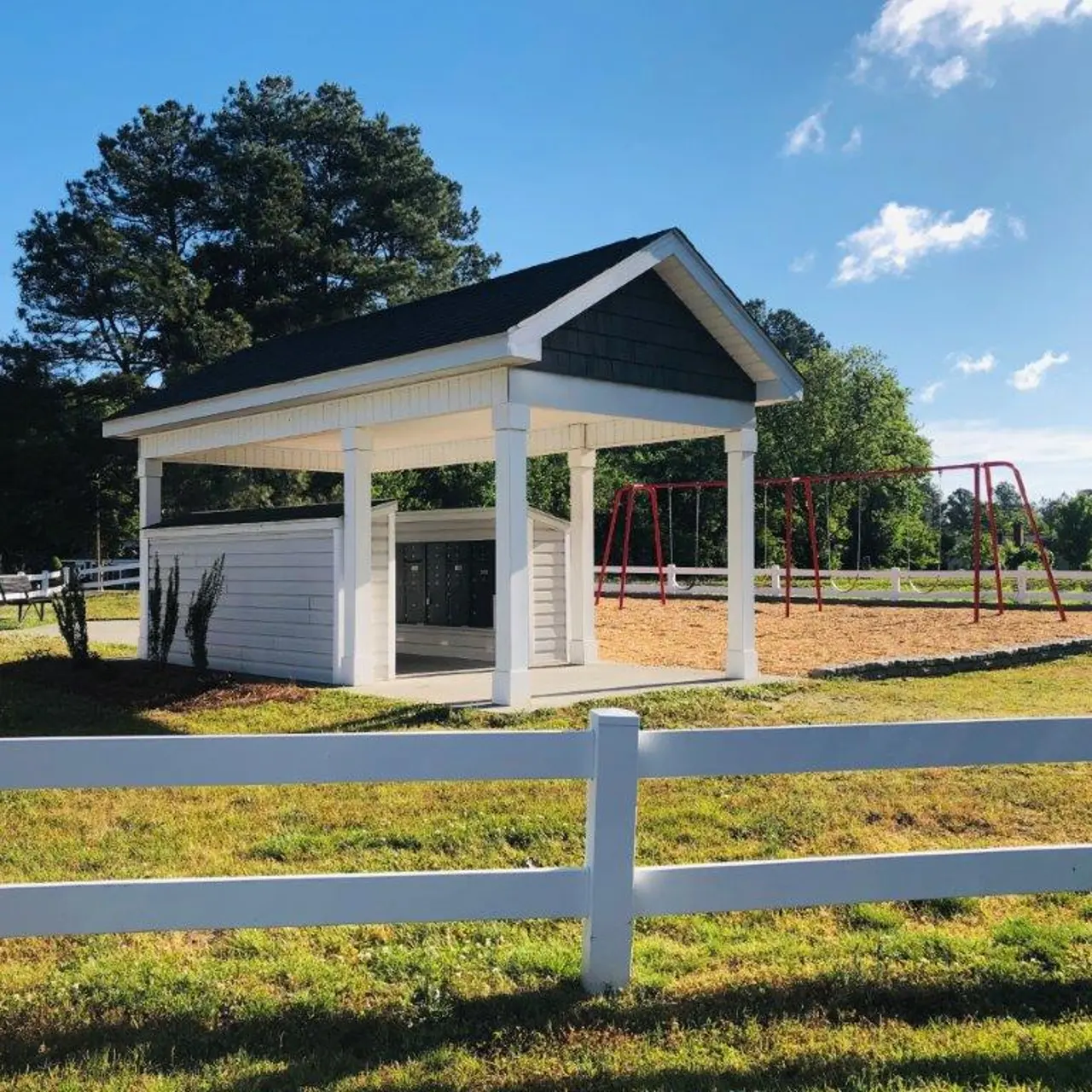 A small white shelter with a black roof near a playground featuring swings, surrounded by a white fence and green grass.