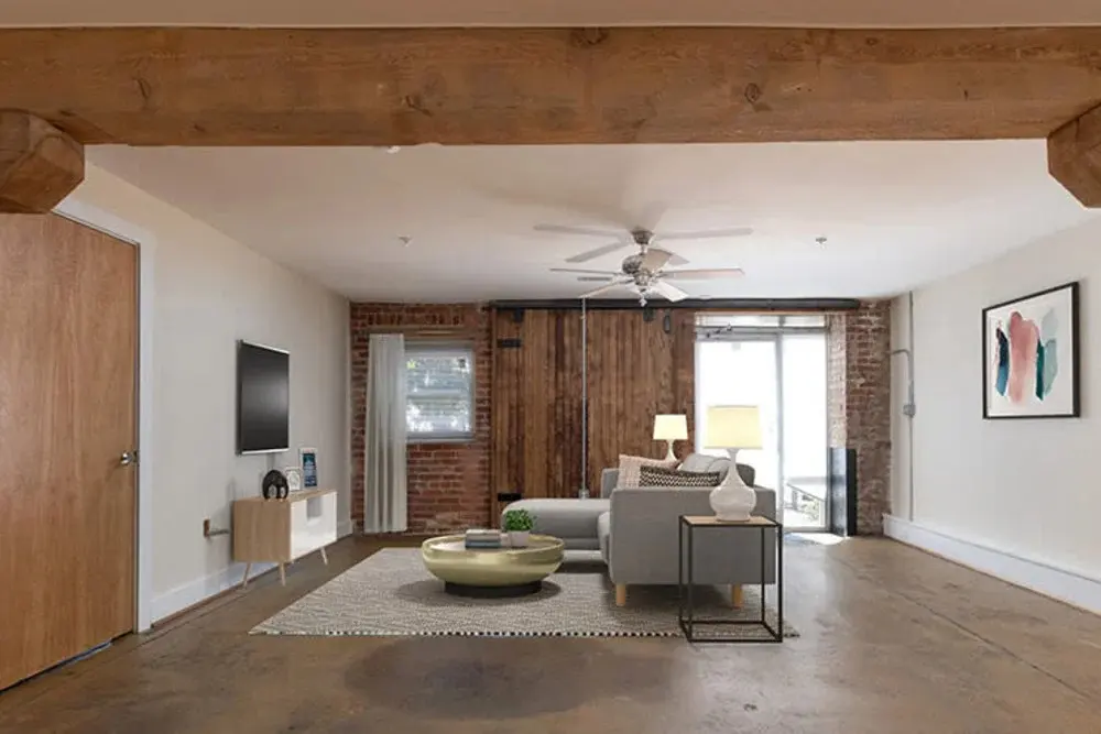 A modern living room featuring a mix of wall textures, including exposed brick and wood accents. There is a light-colored sofa, a coffee table, and a flat-screen TV mounted on the wall. A fan hangs from the ceiling, and large windows provide natural light.