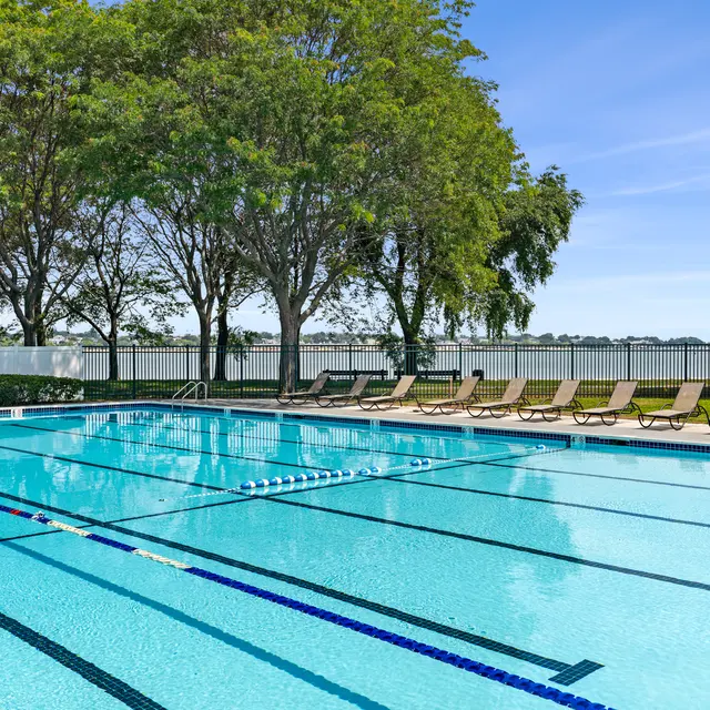 An outdoor swimming pool with lanes marked for swimming, surrounded by green trees and lounge chairs. There is a tranquil lake in the background under a clear blue sky.