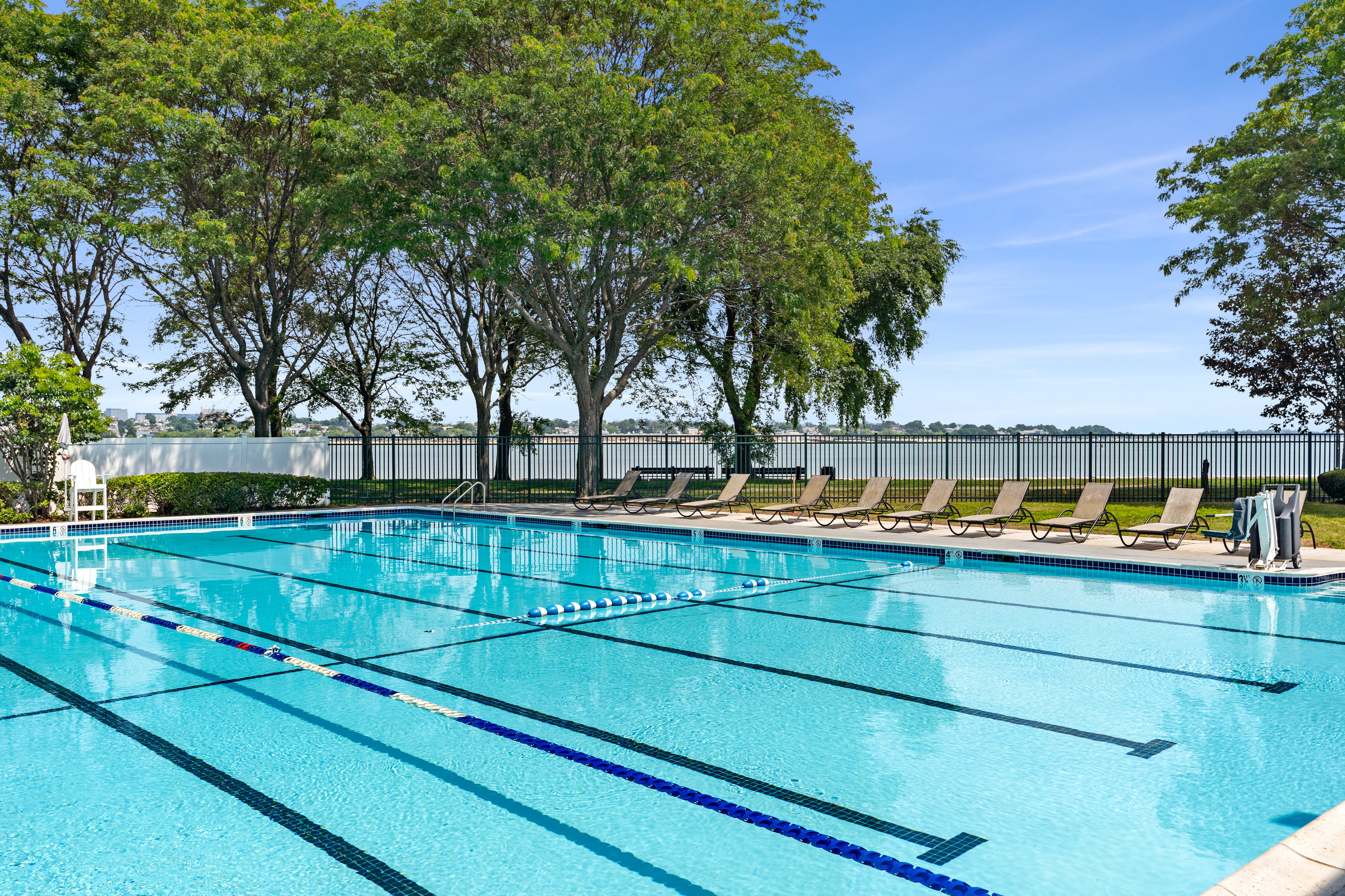 Outdoor Swimming Pool with Lake View An outdoor swimming pool with lanes marked for swimming, surrounded by green trees and lounge chairs. There is a tranquil lake in the background under a clear blue sky.