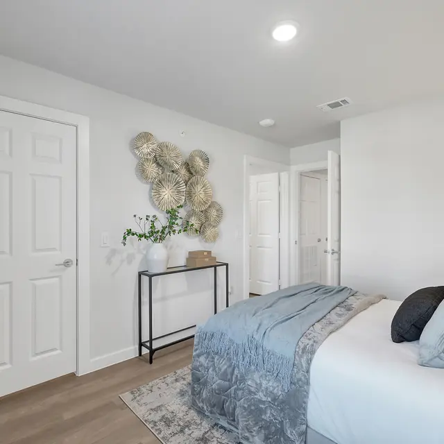 A modern bedroom featuring a neatly made bed with gray and blue bedding, decorative pillows, a stylish chair, and wall art made of natural materials. There is a small table alongside the bed and neutral-colored walls.