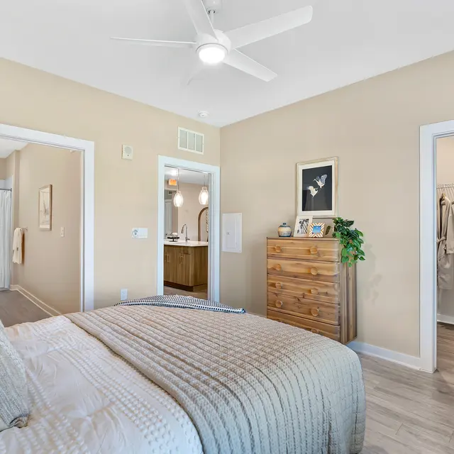 Modern Bedroom Interior A modern bedroom featuring a large bed with a textured blanket, a wooden dresser, and light walls. Doorways to other rooms are visible in the background, along with a ceiling fan and a plant accenting the space.