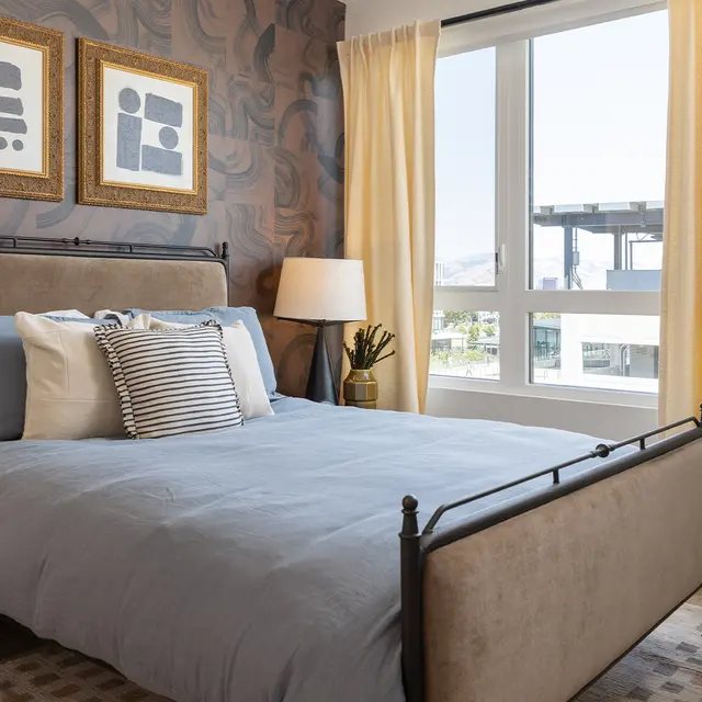 A modern bedroom featuring a queen-sized bed with a light blue duvet, striped and patterned pillows, and framed abstract art on the wall. Two bedside lamps illuminate the room, with sun filtering through large windows draped with sheer yellow curtains. A plant is placed beside the bed, adding a touch of greenery to the space.