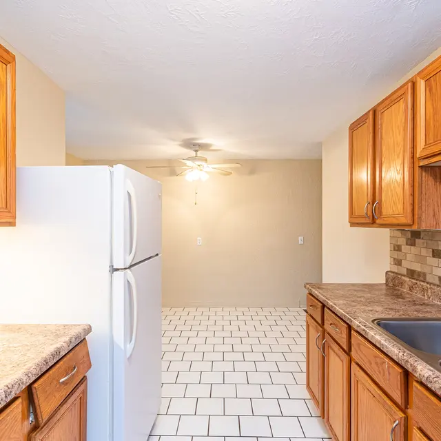 Modern Kitchen Interior A kitchen interior featuring wooden cabinets, a refrigerator, and dual sinks. The flooring is tiled with white squares, and there's a ceiling fan visible in the background.