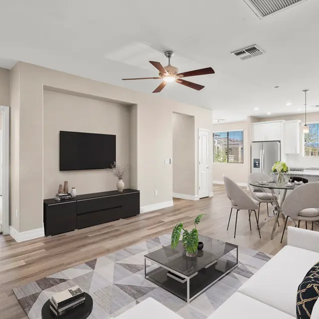 A modern living room featuring a sectional sofa, glass coffee table, and a wall-mounted TV. The space includes a dining area with a round table and chairs, and a kitchen visible in the background. Natural light flows through the windows, creating a bright ambiance. The flooring is wooden, and the walls are painted in a light beige color, enhancing the contemporary feel.