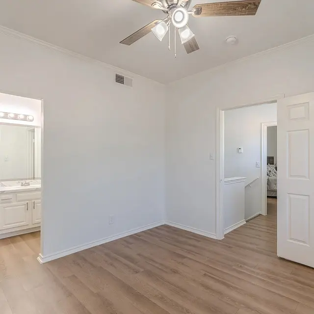 A spacious bedroom with light wood flooring, featuring a ceiling fan and two doorways leading to adjacent rooms. To the left, a bathroom with a double vanity is visible. The walls are painted white, enhancing the bright and airy atmosphere.