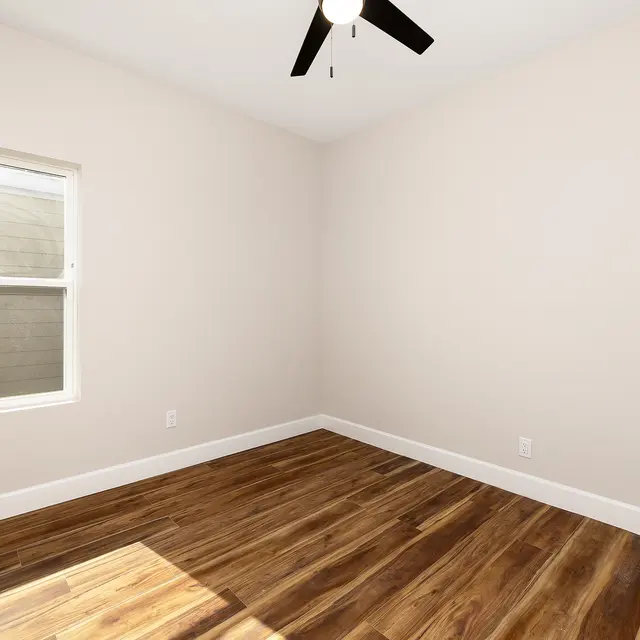 An empty room with light-colored walls and new wooden flooring, featuring a single window and a ceiling fan.