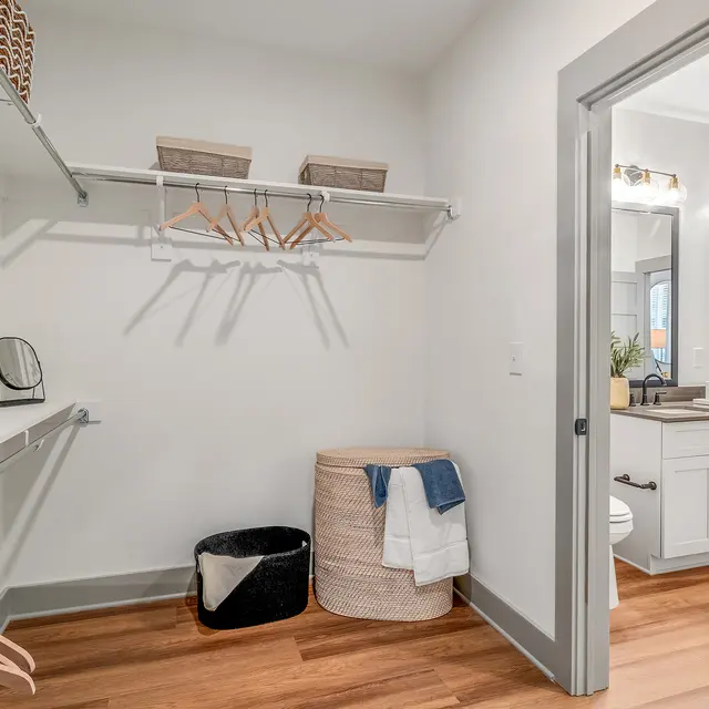 A spacious walk-in closet with wooden flooring, featuring hanging space, storage baskets, and a view into an adjoining bathroom area.