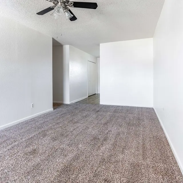 A spacious living room with light gray carpet and white walls. A ceiling fan is visible, and there are doorways leading to other rooms.