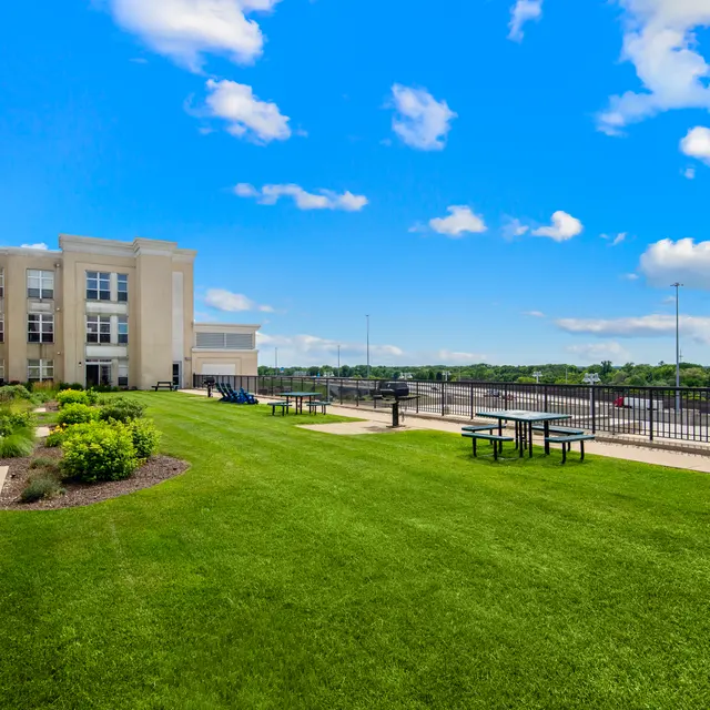 A spacious outdoor lawn area with manicured grass, tables, and a modern building in the background against a bright blue sky with clouds.
