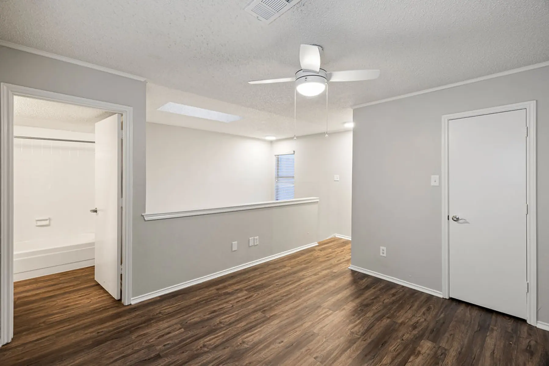 Interior view of an empty room featuring a ceiling fan, light walls, and a wooden floor. There are two doors leading to other spaces, with one showing a bathroom area.