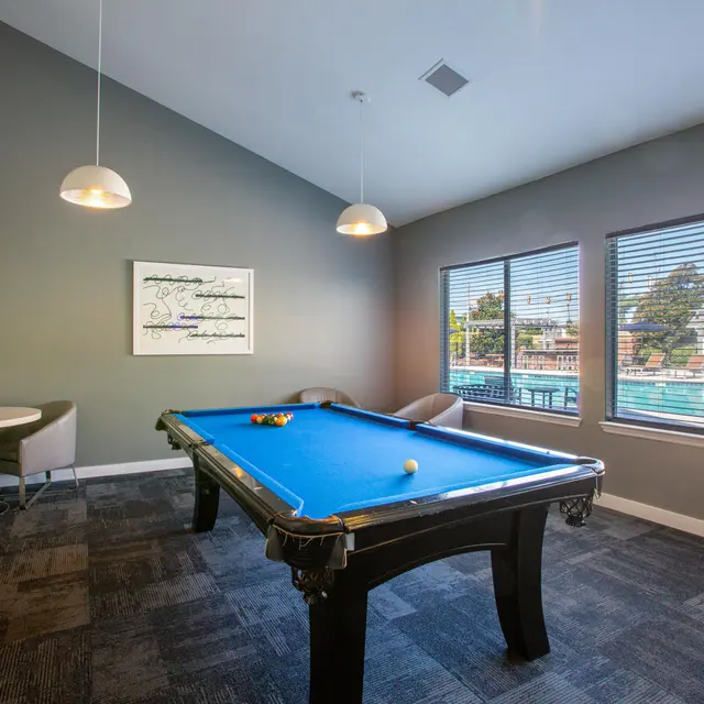 A spacious lounge area featuring a blue pool table, a small round table with chairs, and large windows overlooking a swimming pool. The walls are painted in a soft gray, and a flat-screen TV is mounted on the wall.