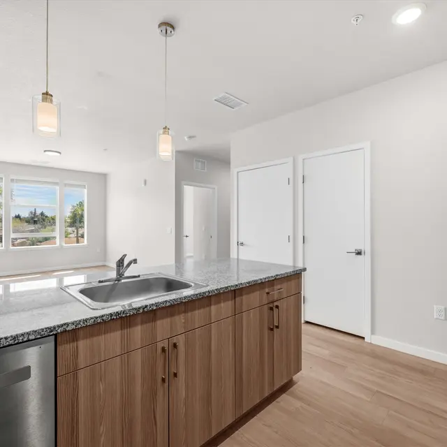 A contemporary kitchen with a granite countertop, stainless steel sink, and wooden cabinetry. Decorative lighting hangs above the counter, and a window provides natural light.
